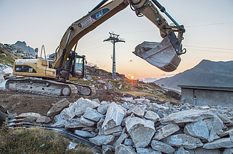 Bagger im Einsatz auf einer Baustelle in hochalpiner Umgebung mit Felsblöcken, Baustelleneinrichtungen und Seilbahninfrastruktur.
