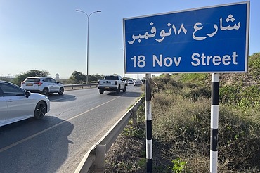 Straßenschild der 18 November Street an einer mehrspurigen Straße mit Fahrzeugverkehr, Leitplanken und begleitender Verkehrsinfrastruktur.ehr