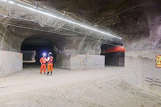 Unterirdisches Bauwerk mit mehreren Stollen, Beleuchtung, Lüftungsleitungen und Versorgungseinrichtungen. Zwei Mitarbeitende in Schutzausrüstung befinden sich im Bereich einer Tunnelverzweigung.