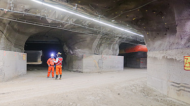 Unterirdisches Bauwerk mit mehreren Stollen, Beleuchtung, Lüftungsleitungen und Versorgungseinrichtungen. Zwei Mitarbeitende in Schutzausrüstung befinden sich im Bereich einer Tunnelverzweigung.