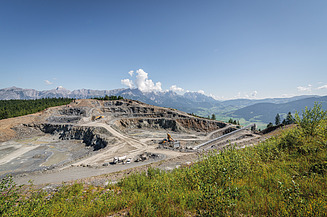 Tagebau mit Abbauflächen, Fördertechnik und Baumaschinen vor einer alpinen Bergkulisse. Sichtbar sind die verschiedenen Gewinnungsstufen des Steinbruchs sowie die umliegende Landschaft.