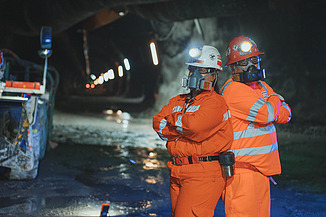 Zwei Mitarbeitende mit Schutzausrüstung in einem unterirdischen Stollen der Mine El Teniente in Chile. Im Hintergrund sind Tunnelinfrastruktur und bergbauliche Anlagen erkennbar.