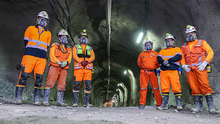 Employees wearing personal protective equipment inside an underground tunnel at Chuquicamata Mine in Chile. Tunnel infrastructure, lighting systems and mining facilities are visible in the background.