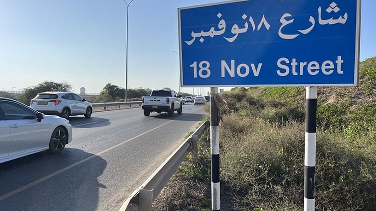 Road sign marking 18 November Street alongside a busy roadway with traffic, guardrails and associated transport infrastructure.