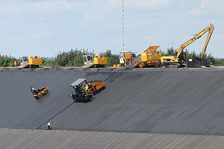Road paving machine laying a new asphalt surface on a transport infrastructure project. The foreground shows the freshly paved asphalt layer and a longitudinal joint.