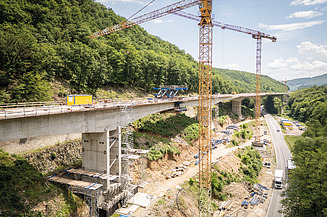 Brückenbaustelle in einem bewaldeten Tal mit Kränen, Betontragwerk und Baustelleneinrichtungen entlang einer Verkehrsverbindung.