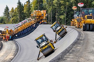 Specialised paving machine and road rollers placing and compacting an asphalt layer on the sloped embankment of a dam or reservoir structure.