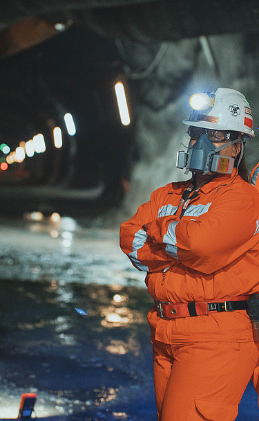 Zwei Mitarbeitende mit Schutzausrüstung in einem unterirdischen Stollen der Mine El Teniente in Chile. Im Hintergrund sind Tunnelinfrastruktur und bergbauliche Anlagen erkennbar.