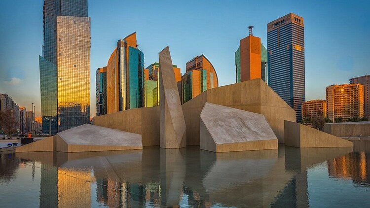 Architectural monument in Abu Dhabi surrounded by a reflecting pool, with contemporary high-rise buildings forming the city skyline in the background.