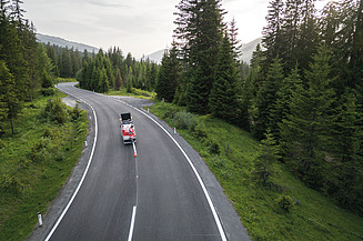 Aerial view of a winding road passing through a forested alpine landscape, with a service vehicle carrying out survey, inspection or maintenance activities.