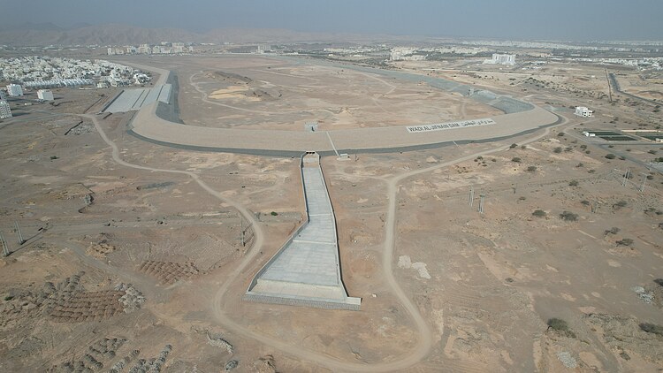 Aerial view of the Wadi Al-Jifnain flood protection dam in an arid landscape, showing the dam structure, reservoir area and surrounding terrain.